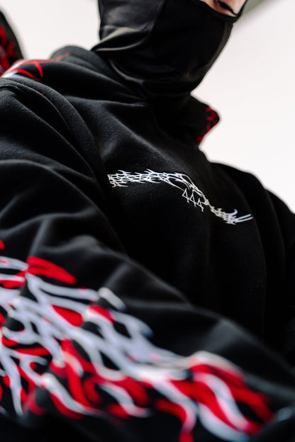 Detail shot of the Reflective Hoodie v2 chest, showing the white 444 mark in tribal script on jet-black fleece, with the red and white sleeve print below.