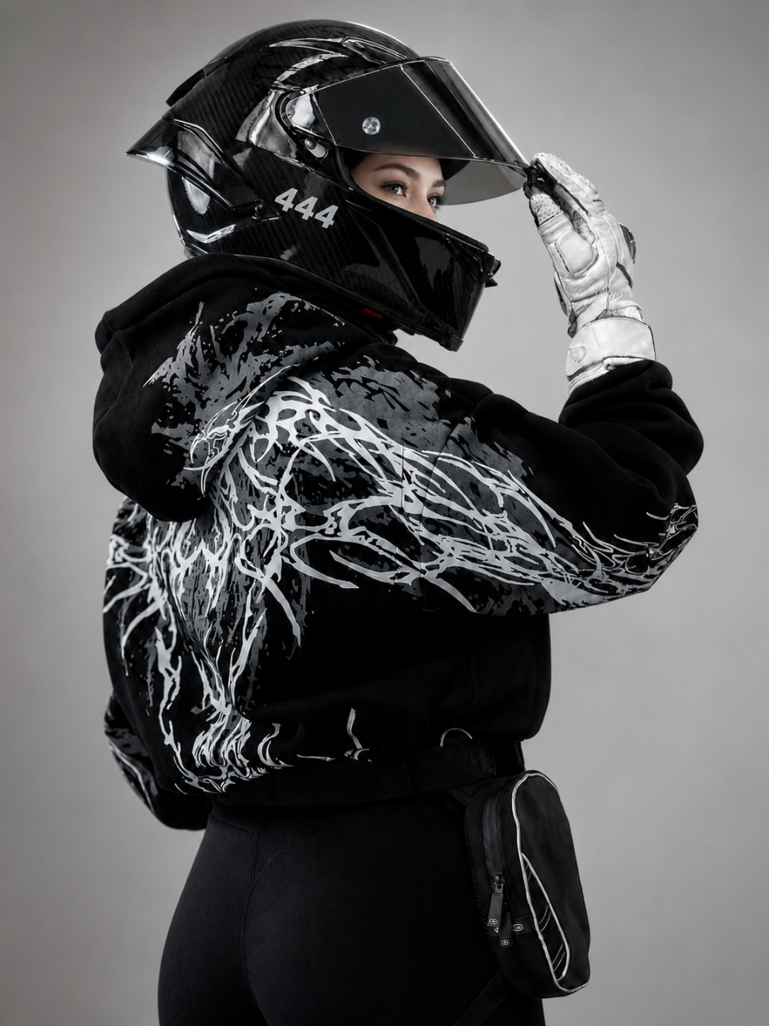 A person wearing a black full-face racing helmet with a clear visor, a black hooded jacket with a bold white abstract graphic print across the back and sleeves, a white racing glove, and black pants, posed against a plain gray studio background.