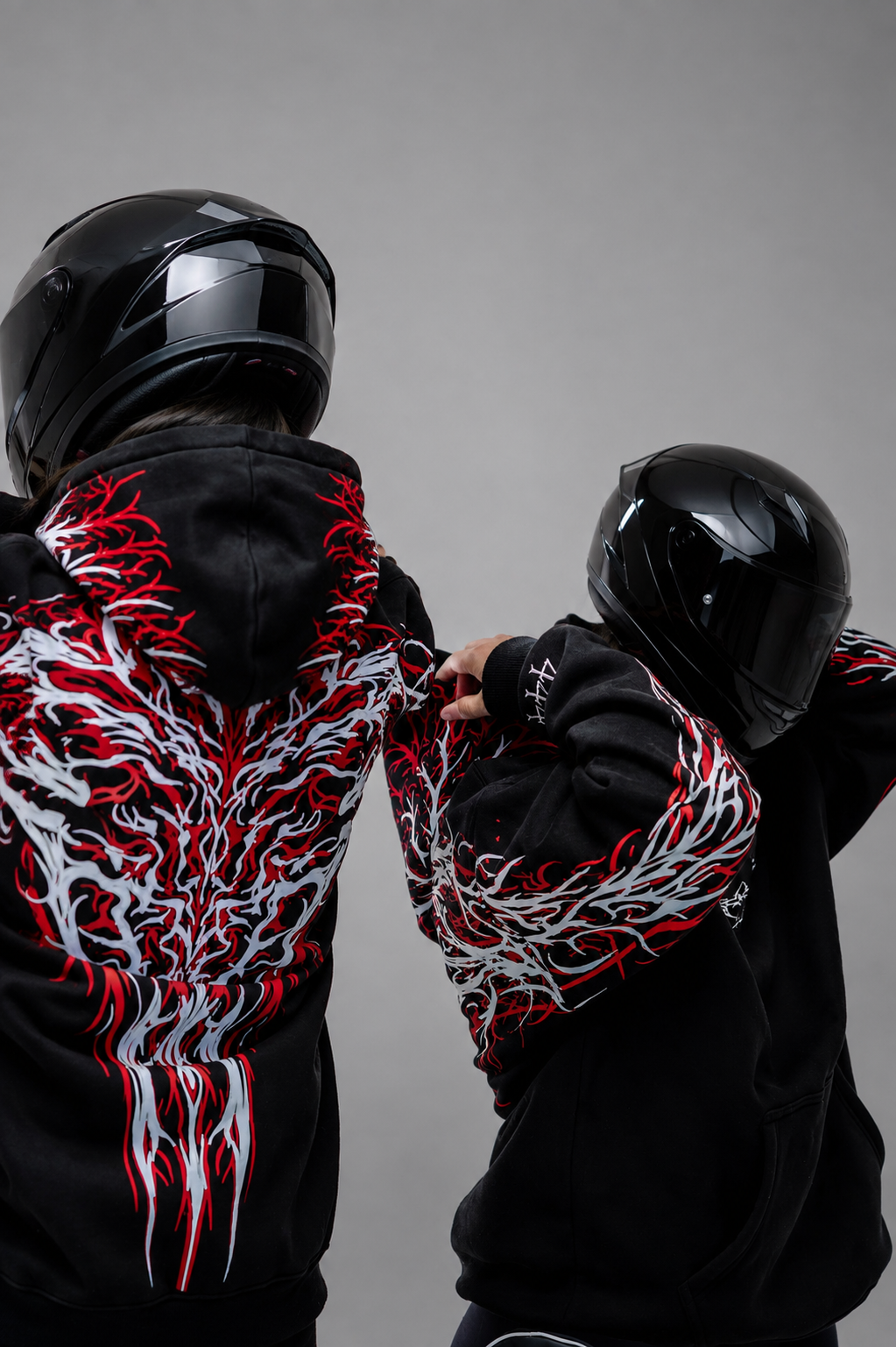 Two riders in black full-face helmets seen from behind, both wearing the Reflective Hoodie v2. The red and white tribal print covers the backs and sleeves, against a plain grey studio background.