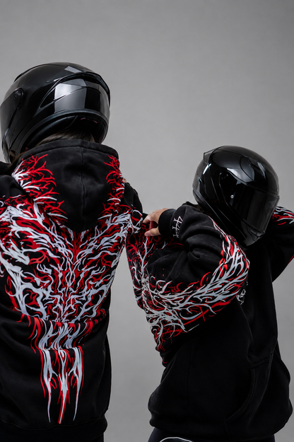 Two riders in black full-face helmets seen from behind, both wearing the Reflective Hoodie v2. The red and white tribal print covers the backs and sleeves, against a plain grey studio background.