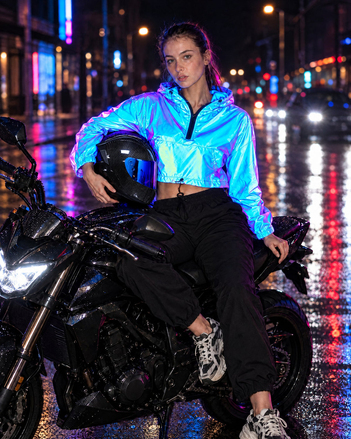 Woman in a Cobalt reflective cropped jacket sitting on a motorcycle at night, holding a carbon-look full-face helmet