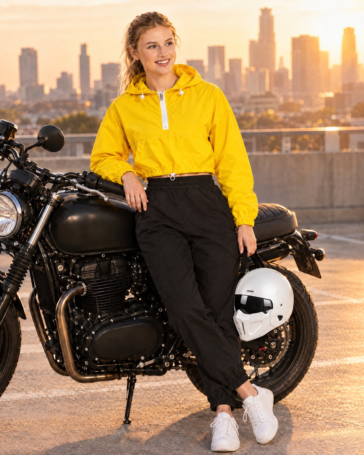 Woman in a Gold yellow cropped hooded jacket leaning on a motorcycle at golden hour with a white convertible helmet